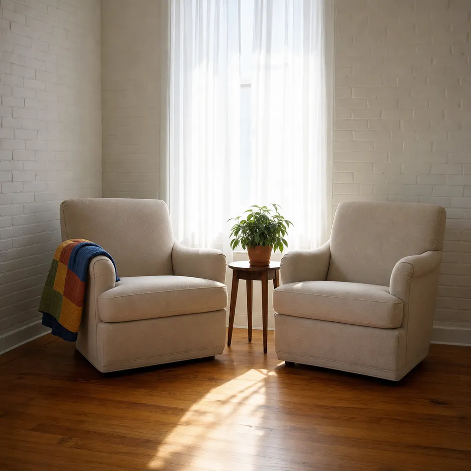 An empty armchair with a patchwork throw beside a bright window, and a plant on a small side table.