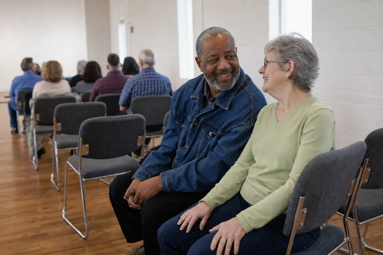 Two older friends smile at each other in a row of chairs, with other people seated in the rows behind them.