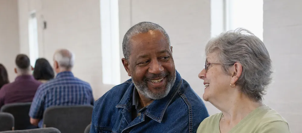 Two older friends smile at each other in a row of chairs, with other people seated in the rows behind them.